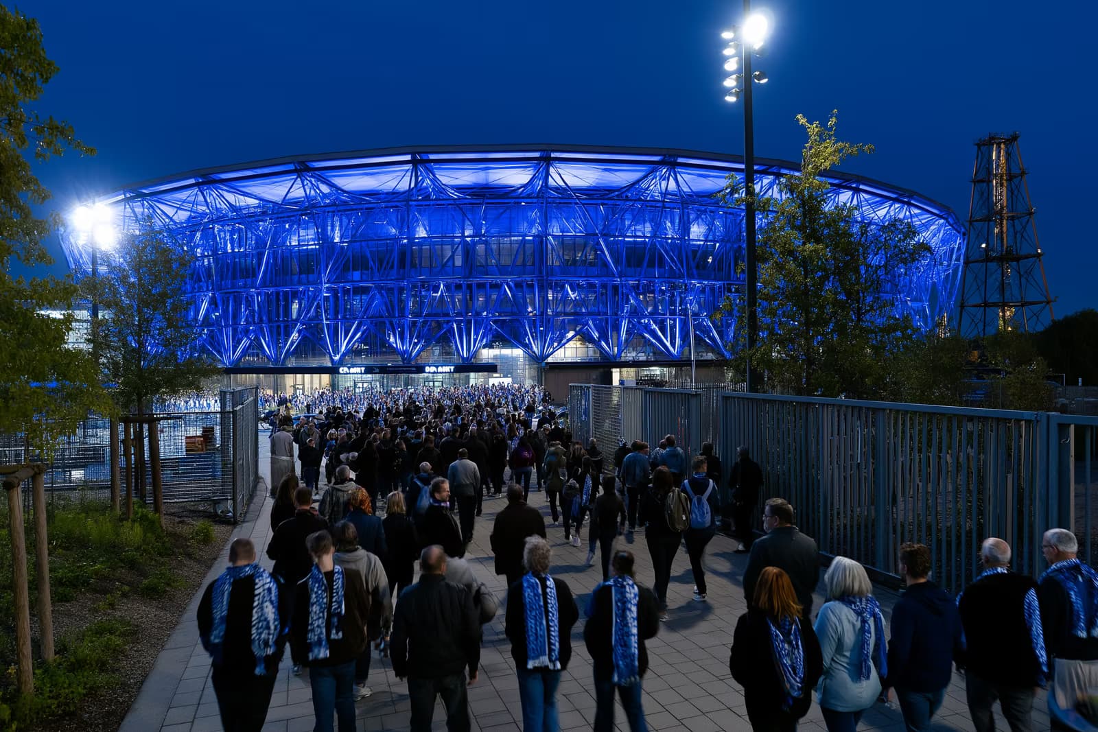 Uno stadio di calcio nella regione della Ruhr si tinge di un blu reale di notte, mentre i tifosi con le sciarpe si riversano verso i cancelli dopo il trasferimento del Gosens Schalke.