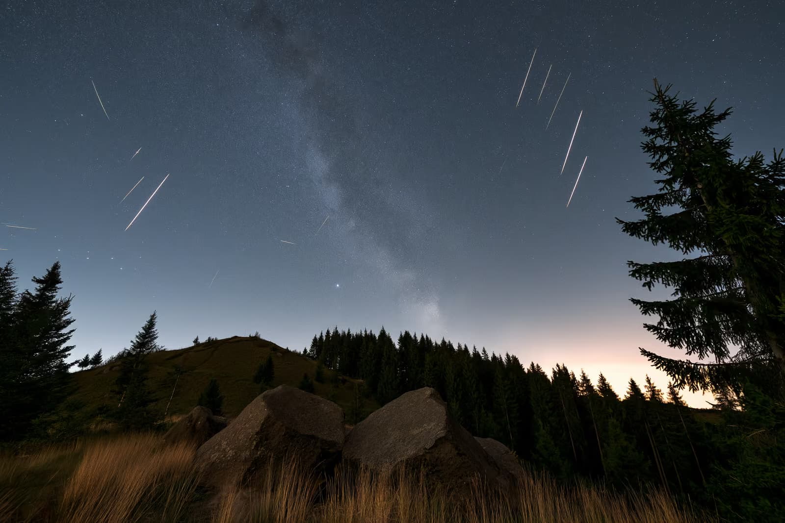 Un cielo notturno sopra una buia campagna tedesca, con scie meteoriche e una debole fascia di stelle in alto.